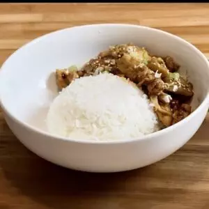 White rice with flavorful stir-fried chicken and vegetables in a white bowl on a wooden table.